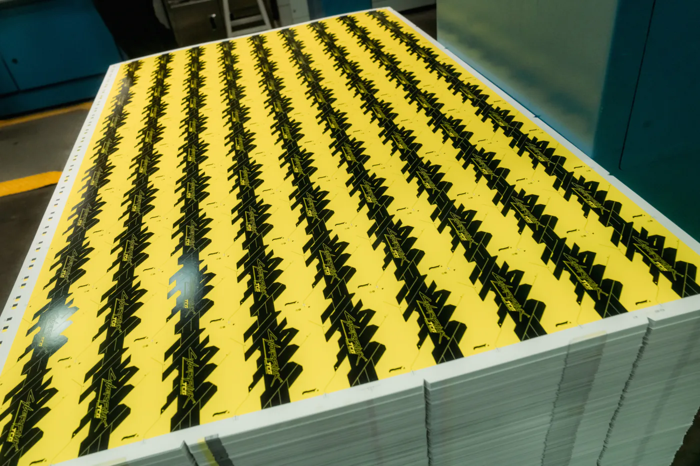 An uncut press sheet of Cyberpunk TCG card backs - rows of the yellow-and-black Cyberpunk TCG logo - sitting on top of a tall stack of printed sheets inside the RR Donnelley print facility.