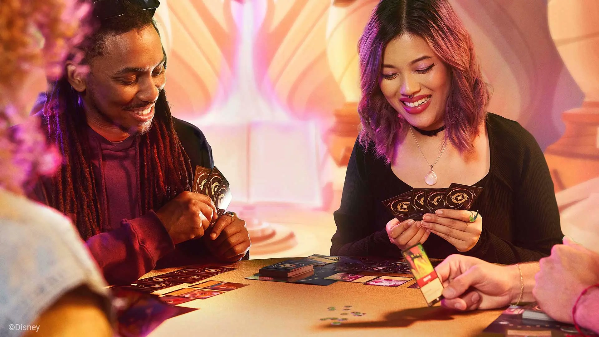 Four players around a table mid-game in a multiplayer Disney Lorcana match, two of them laughing over fanned hands of cards, in the official promotional photo for the Format Coconut announcement.