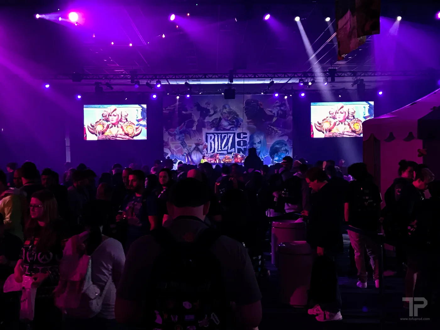 A packed BlizzCon crowd facing a stage under purple lighting, with the BlizzCon logo and character art on a backdrop screen and two side screens showing panel footage.