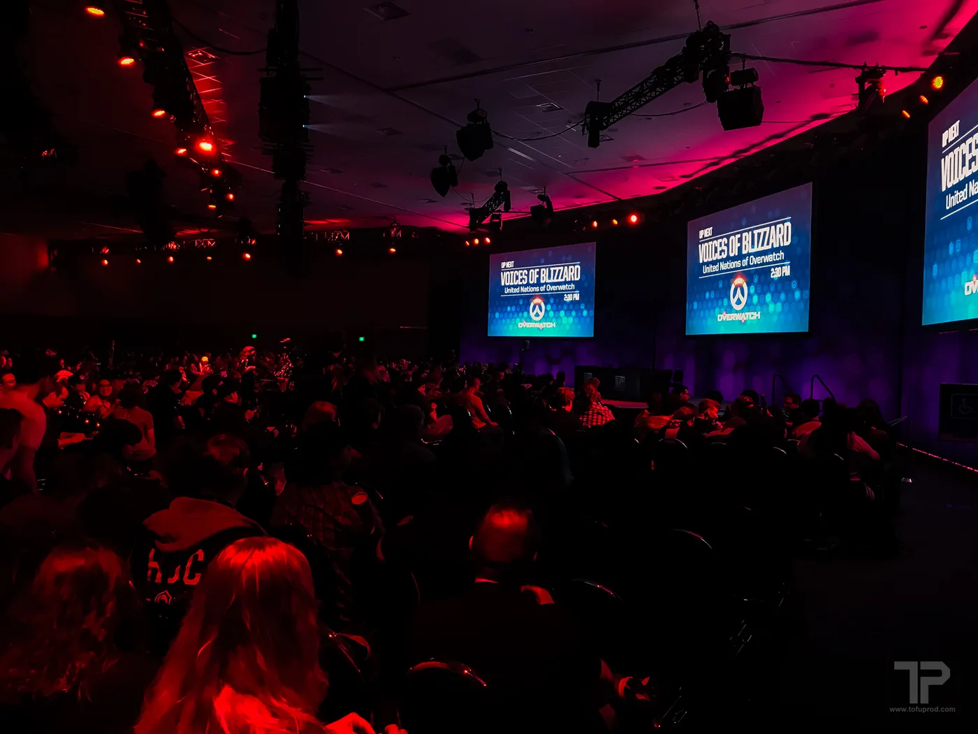 A crowd facing large screens reading "UP NEXT: VOICES OF BLIZZARD — United Nations of Overwatch, 2:30 PM" under red stage lighting at a BlizzCon panel hall.