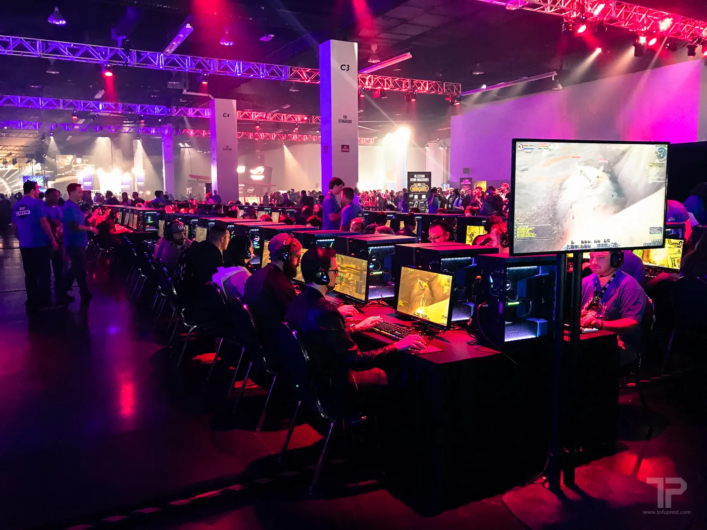 People playing World of Warcraft on rows of demo PCs at BlizzCon, watched by a crowd, with a large overhead screen also showing a WoW gameplay feed.