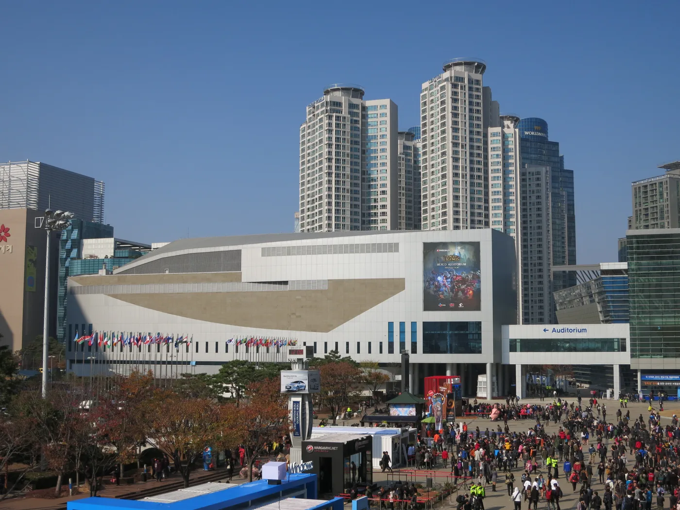 BEXCO Auditorium in Busan seen across the plaza on a clear autumn day: a long, low white and beige building with a row of national flags along its front, a large League of Legends poster reading BEXCO AUDITORIUM 2013.11.15 - 11.16 on its facade, an Auditorium sign over the entrance on the right, high-rise towers behind, and a dense crowd of visitors on the plaza below.