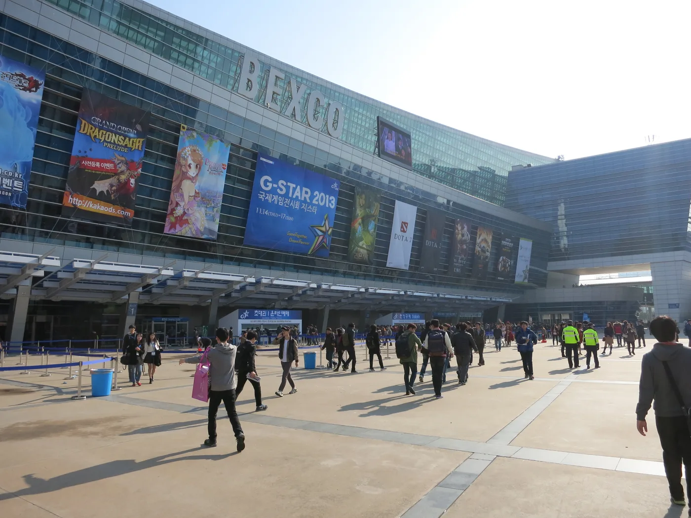 The glass front of BEXCO Exhibition Center 1 in Busan on a sunny day, with BEXCO in large letters along the roofline above a row of hanging game banners, the centre one a blue G-STAR 2013 banner with a multicoloured star, while visitors with backpacks cross the paved plaza and staff in yellow high-visibility vests stand near the entrance canopy.