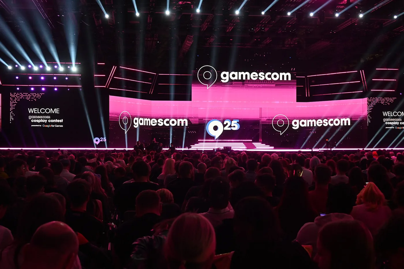 A packed arena audience seen from behind, facing a huge stage lit in hot pink with three illuminated gamescom logos and a screen reading WELCOME gamescom cosplay contest, white spotlight beams crossing above the crowd.