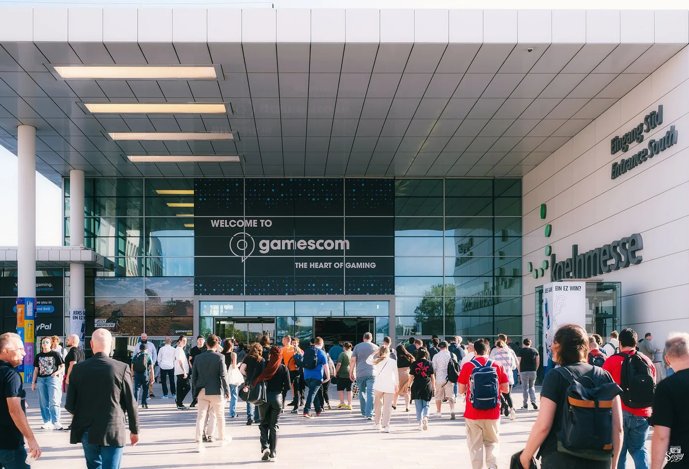 The south entrance of Koelnmesse during gamescom: a wide white canopy over a glass facade carrying a large banner reading WELCOME TO gamescom THE HEART OF GAMING, with koelnmesse and Eingang Sued / Entrance South lettering on the wall at right and a crowd of visitors with backpacks walking toward the doors in bright sunlight.