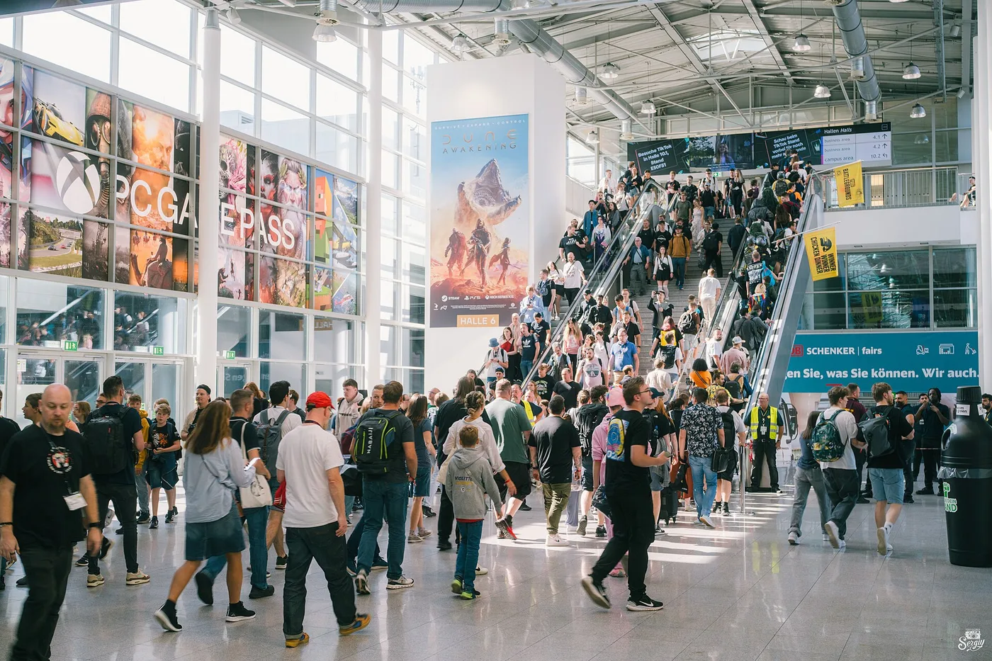 A packed glass-walled concourse at Koelnmesse during gamescom: hundreds of visitors with backpacks stream across a bright tiled floor while two escalators carry more people down past a Dune Awakening banner marked HALLE 6, with a PC Game Pass photo wall along the window and a hall directory sign listing business area 4.1, business area 4.2 and campus area 10.2.