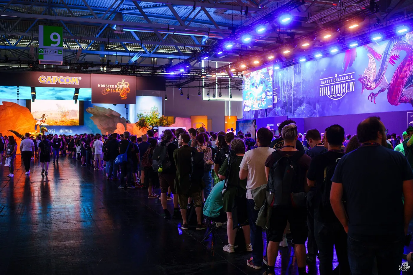 A long queue of visitors with backpacks stretching down a dark exhibition hall at gamescom, lit purple and blue, past a Capcom and Monster Hunter Wilds booth on the left and a Final Fantasy XIV Battle Challenge video wall on the right, with a green hall marker numbered 9 overhead.
