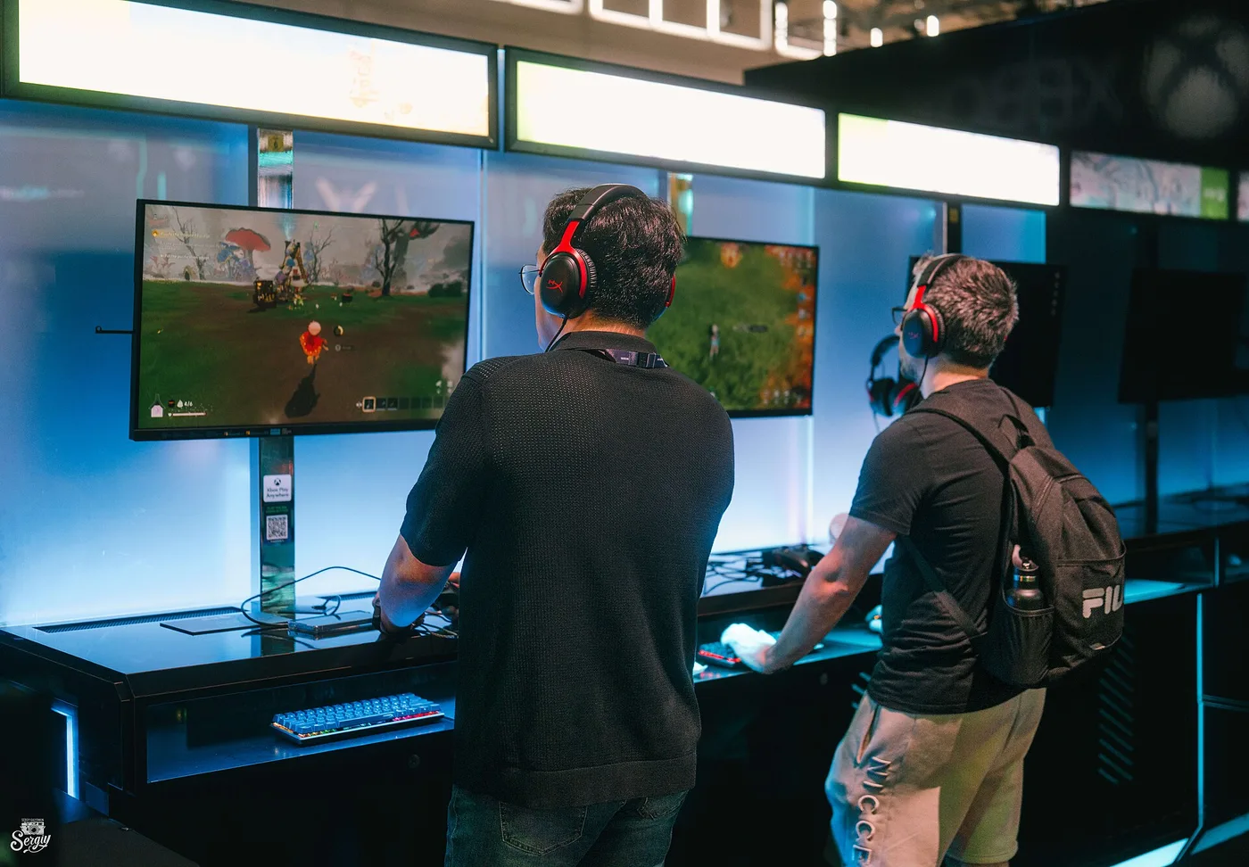 Two visitors in headsets standing at a bank of demo stations at gamescom, playing on large monitors mounted above a black counter with mechanical keyboards, lit in blue with an Xbox logo faintly visible on the wall behind.