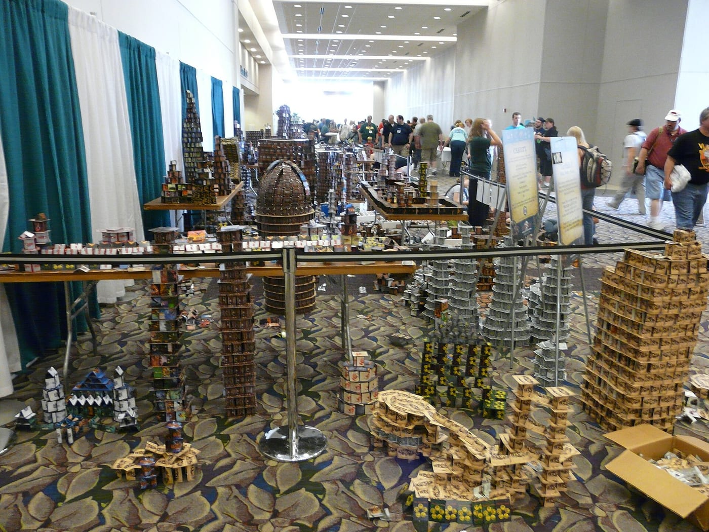 A convention hall carpet covered in dozens of towers, walls and a domed structure built entirely from stacked and folded trading cards, roped off behind retractable belt barriers, with a printed CHARITY sign beside the display and attendees walking past in the background.
