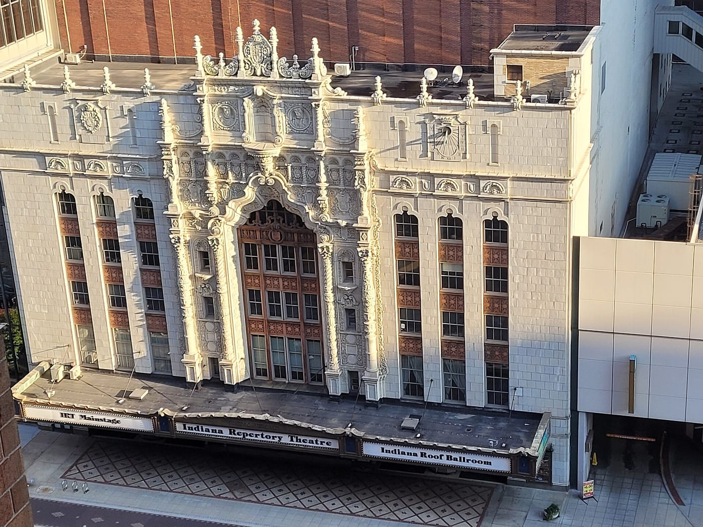 An elevated view of the Indiana Repertory Theatre in downtown Indianapolis: an ornate cream-white Spanish Baroque terra cotta facade with a carved central arch, medallions and finials along the roofline, above a black marquee reading IRT Mainstage, Indiana Repertory Theatre and Indiana Roof Ballroom.