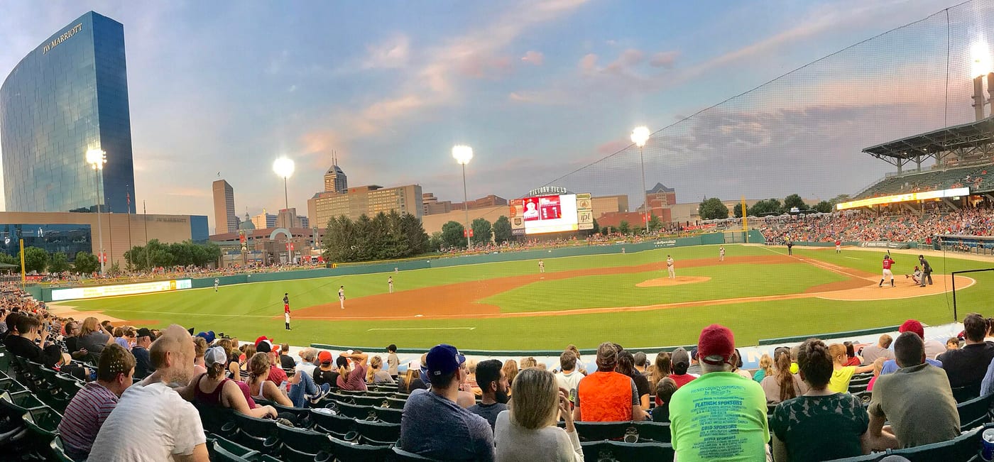A sunset panorama from the seats at Victory Field in Indianapolis, looking across the infield toward the downtown skyline and the curved dark-glass JW Marriott tower, with a full crowd in the stands and the scoreboard reading Victory Field.