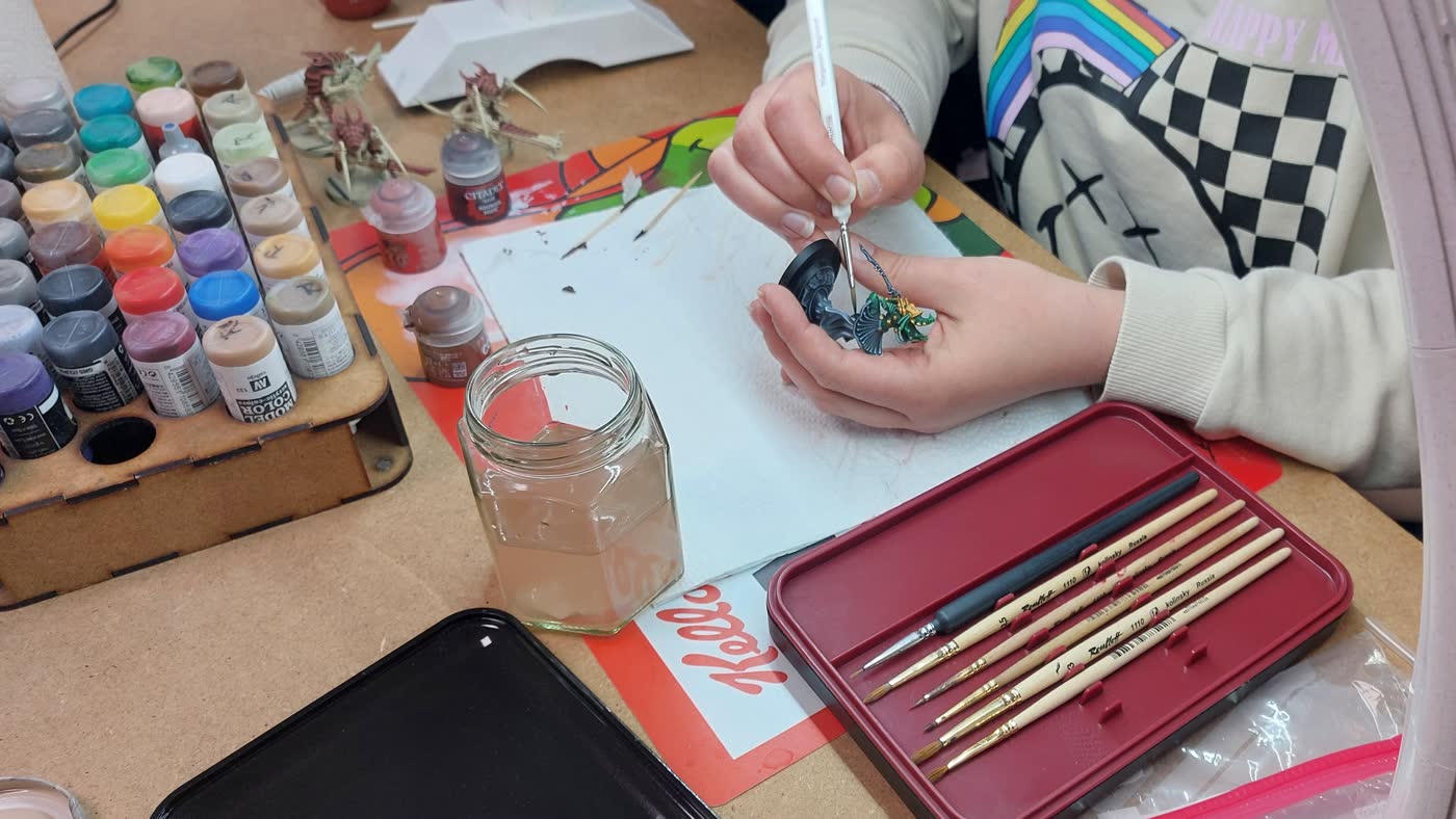 A close-up of a person holding a part-painted miniature between finger and thumb over a hobby desk, applying teal and gold detail with a fine brush, beside a wooden rack of Citadel and Vallejo paint pots, a glass jar of murky brush water, a paper towel and a red tray holding five detail brushes.