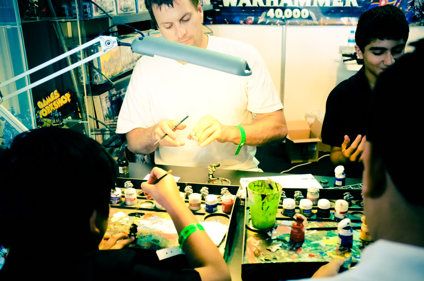 A Games Workshop convention painting stand: a man in a white shirt stands behind an angled desk lamp holding a fine brush over a part-painted miniature on a pot, while seated attendees in the foreground paint their own models on spattered palettes surrounded by rows of Citadel paint pots, a green water cup and a rack of brushes, with a Warhammer 40,000 banner and a Games Workshop display cabinet behind him.