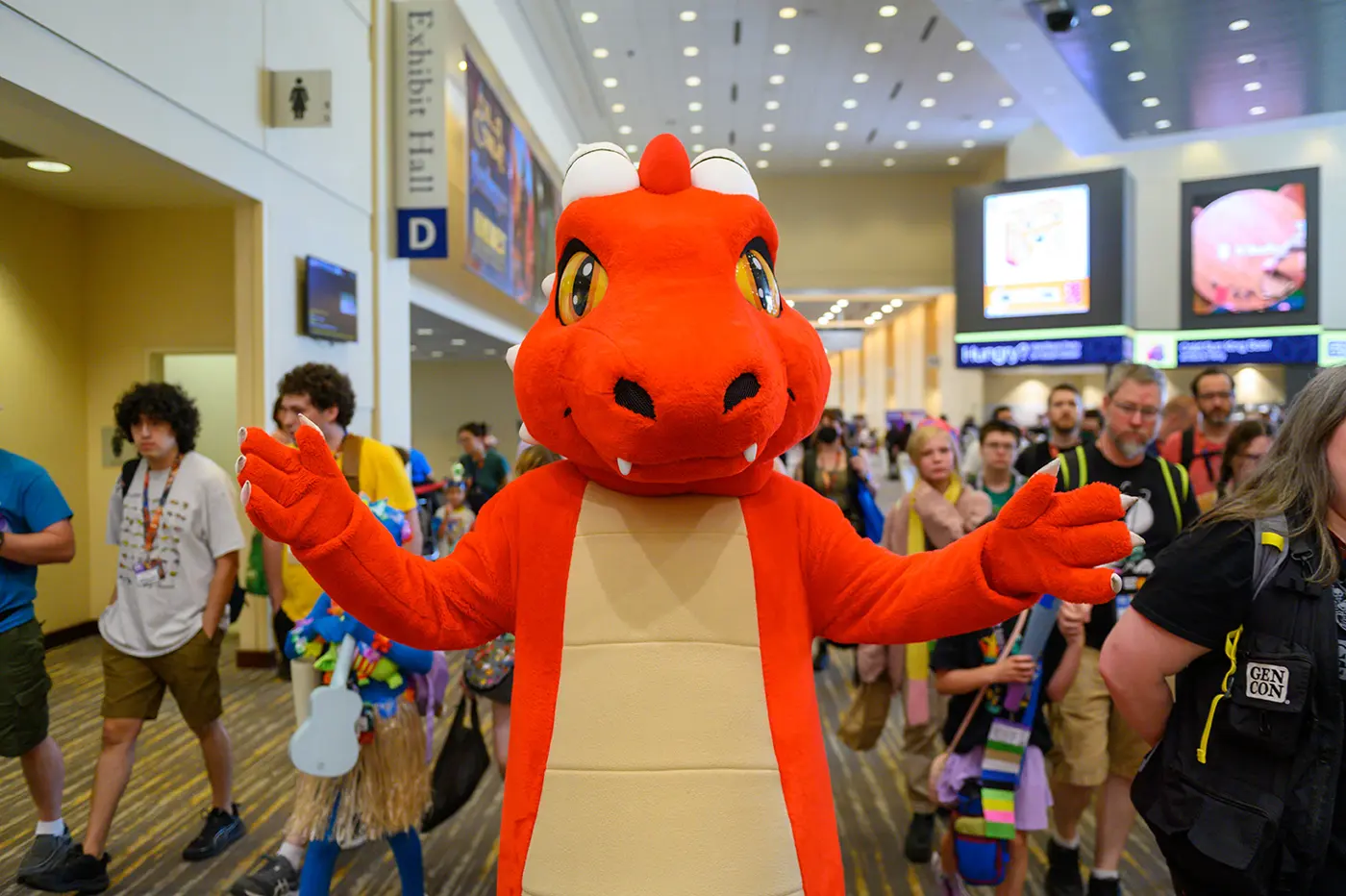 A red plush dragon mascot with outstretched claws standing in a packed Indiana Convention Center concourse at Gen Con, under a blue Exhibit Hall D sign, surrounded by badge-wearing attendees with backpacks and a Gen Con-branded bag.