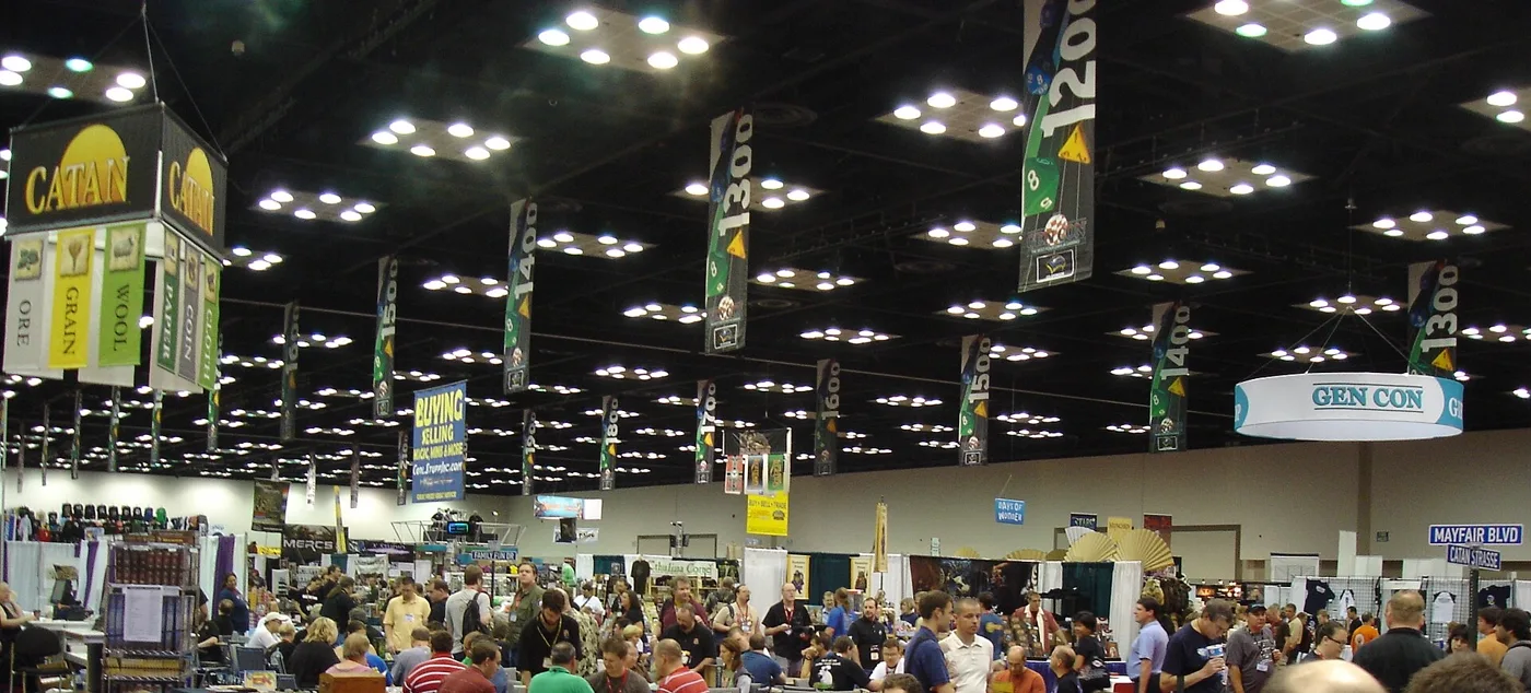 A wide view across the Gen Con exhibit hall in the Indiana Convention Center: numbered aisle banners reading 1200, 1300, 1400, 1500 and 1600 hanging from a dark exposed ceiling grid full of downlights, a circular GEN CON sign at right, a Catan booth banner at left, and a dense crowd of attendees moving between booths.