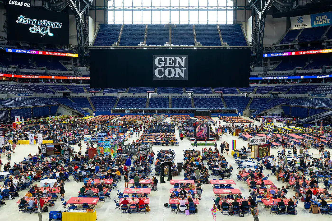 The floor of Lucas Oil Stadium converted into a Gen Con gaming hall — hundreds of red and blue folding tables filled with players, ringed by empty stadium seating, under a huge black banner reading GEN CON and ribbon boards reading "Welcome Gamers!" and "The Best Four Days In Gaming!"
