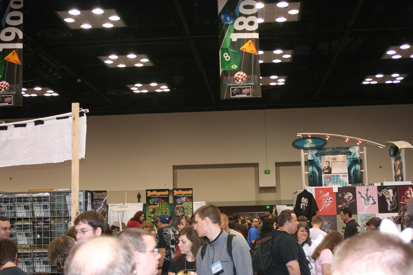 A packed Gen Con exhibit hall seen over the heads of the crowd, with tall banners hanging from the black ceiling grid showing oversized polyhedral dice, an aisle marker reading 180, and a GEN CON banner captioned The Best Four Days In Gaming.