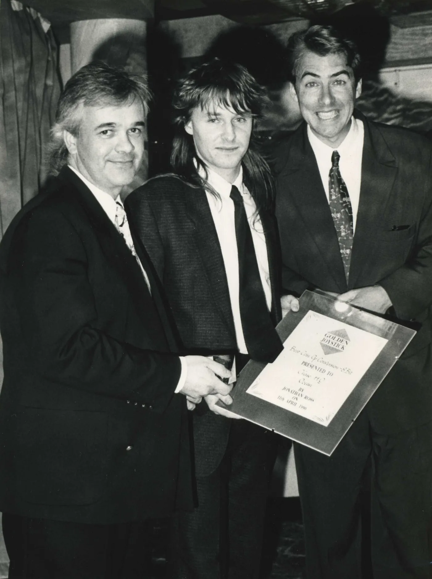 A black-and-white photograph from the 1990 Golden Joystick Awards: three men in suits and ties posing close together, holding a framed certificate with a Golden Joystick diamond logo that reads Best Coin Op Conversion 8 Bit, presented to Chase HQ, Ocean, by Jonathan Ross on 11th April 1990.