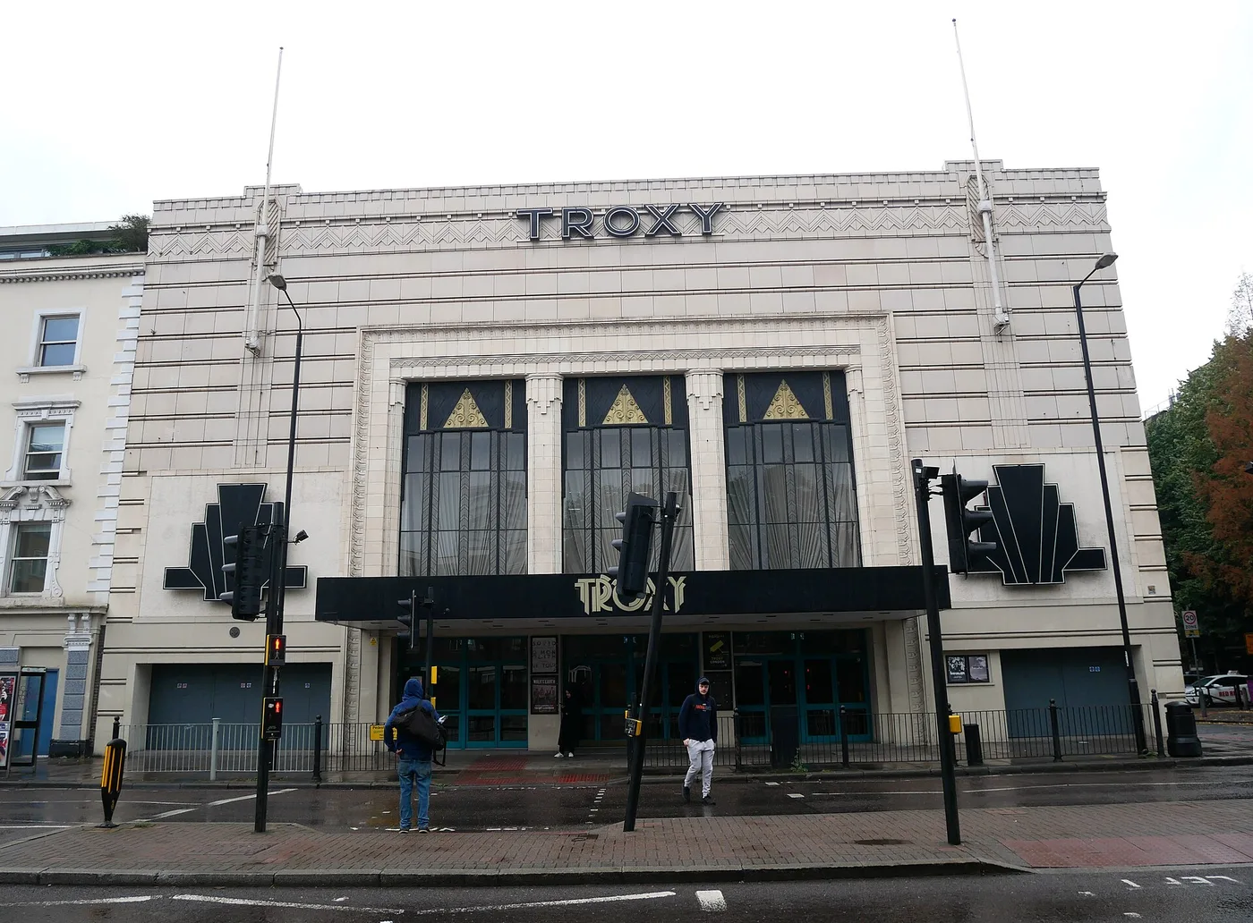 The white art deco front of the Troxy on Commercial Road in Limehouse, east London, on an overcast day: TROXY in dark capitals across the top of the facade and again in gold art deco lettering on the black canopy over the entrance, three tall windows topped with gold triangles, a black fan-shaped ornament on each side, traffic lights and railings along a wet pavement, and two pedestrians on the pavement.
