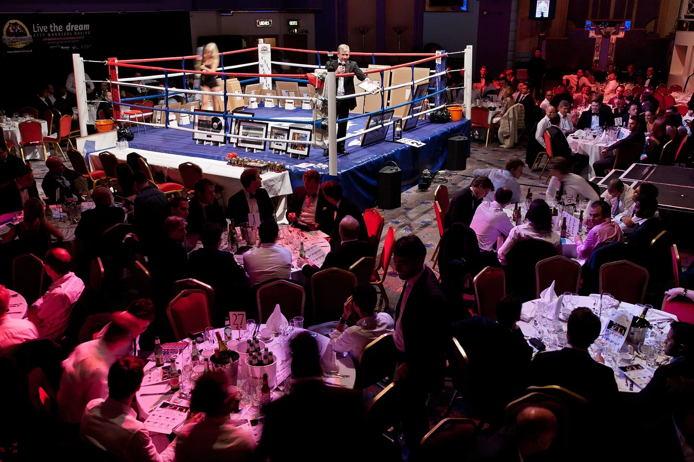The Troxy's main hall set up for a charity auction in 2010: guests in black tie seated at round tables with white cloths, wine bottles and red-backed chairs, gathered around a boxing ring on a raised blue platform where a man in a dinner jacket holds a microphone among framed pictures and records propped up for sale, with more tables filling the floor behind.
