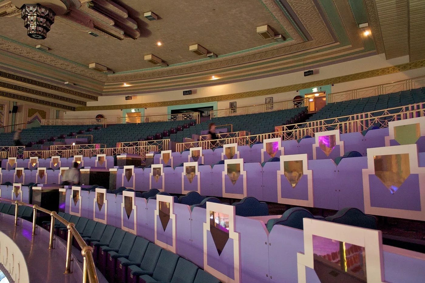The Troxy's upper stalls, almost empty during a 2011 open day: rows of dark teal seats behind tiers of lilac panels, each inset with a pointed gold art deco motif, a brass handrail running along the front, a stepped ceiling with recessed lights, and a geometric chandelier hanging at top left.