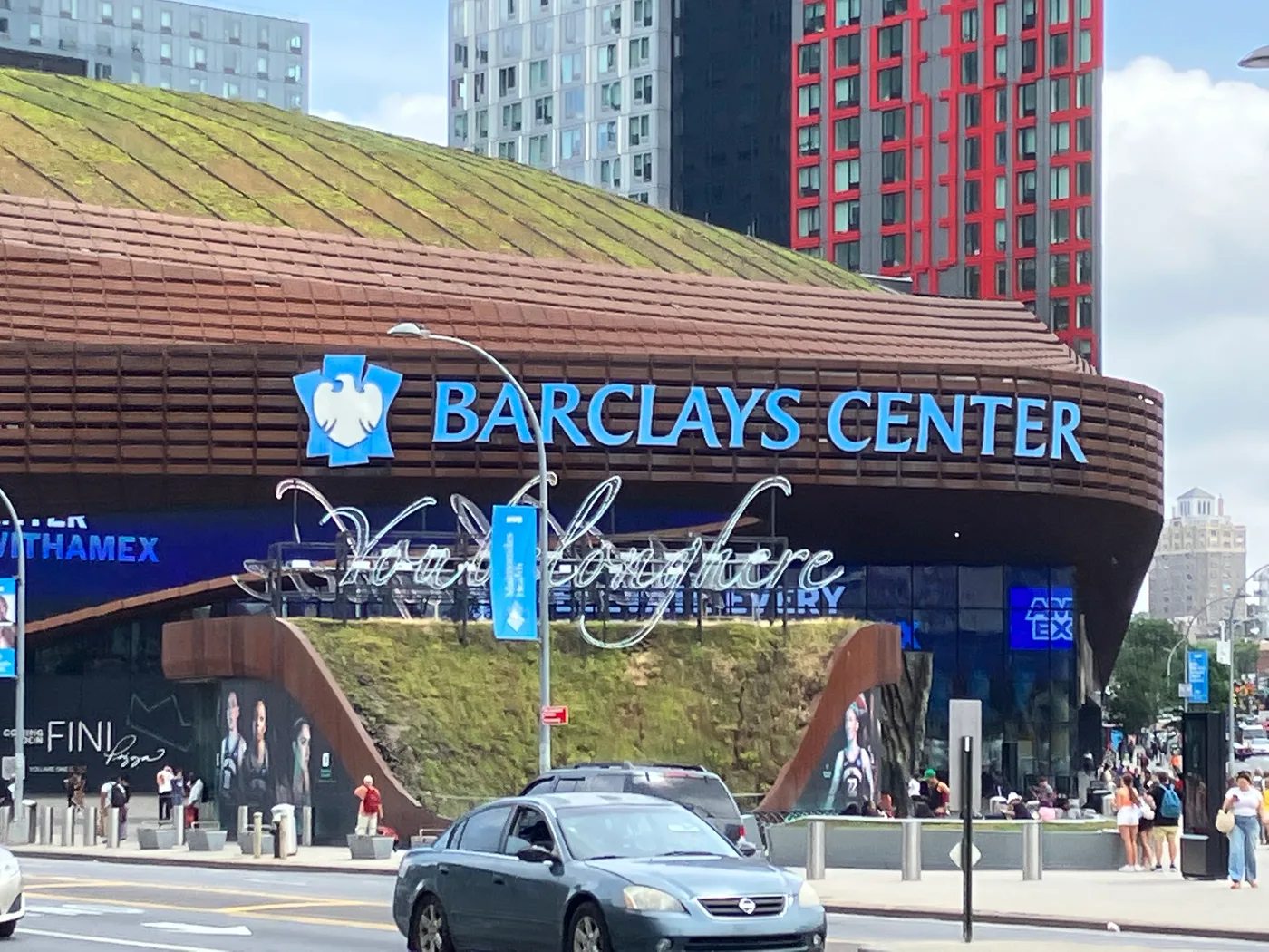 The weathered-steel and glass facade of Barclays Center in Brooklyn, photographed from street level, its curved canopy entrance and illuminated signage visible against the surrounding buildings.