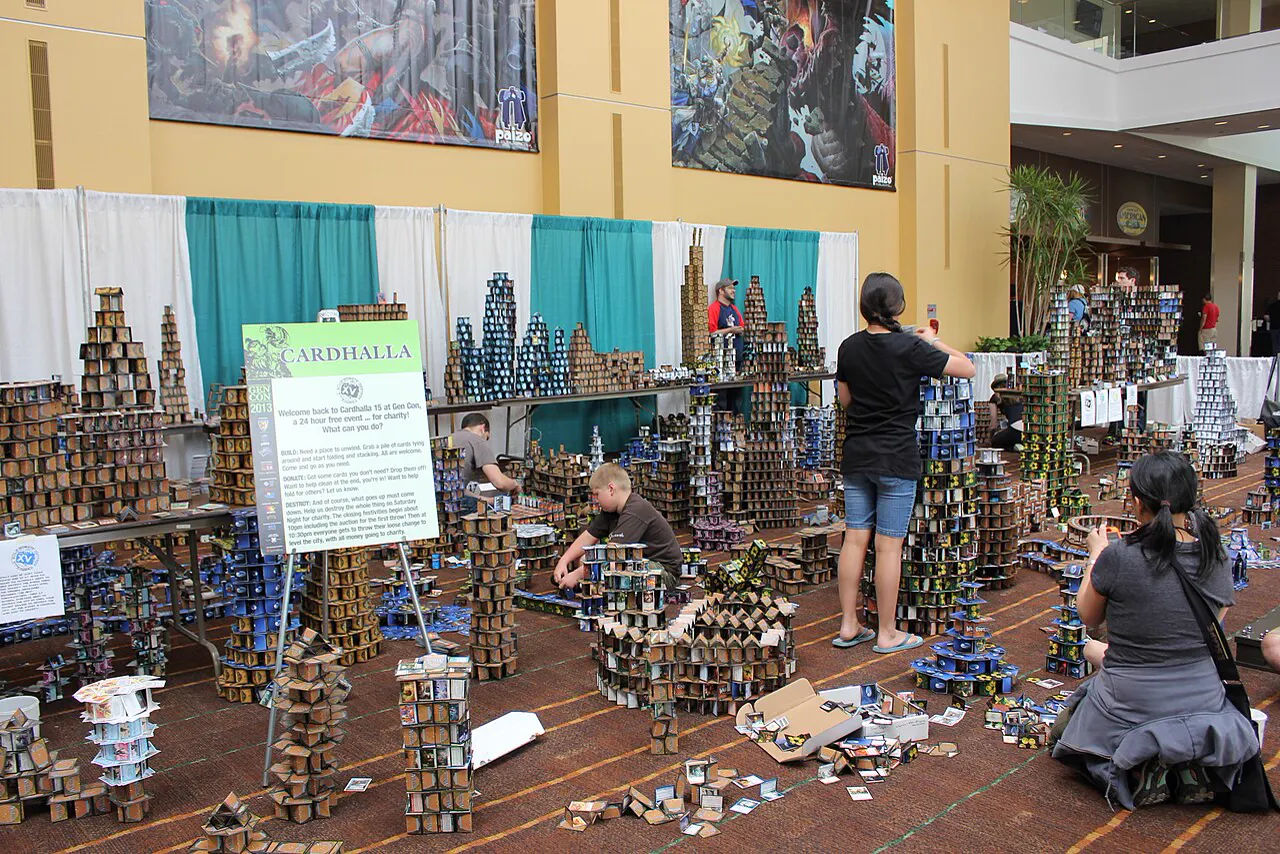 A convention hall lobby at Gen Con with dozens of towers, walls and low structures built from stacked and folded trading cards spread across the carpet, several people crouched adding pieces, a printed CARDHALLA sign on an easel to the left and large hanging art banners above.