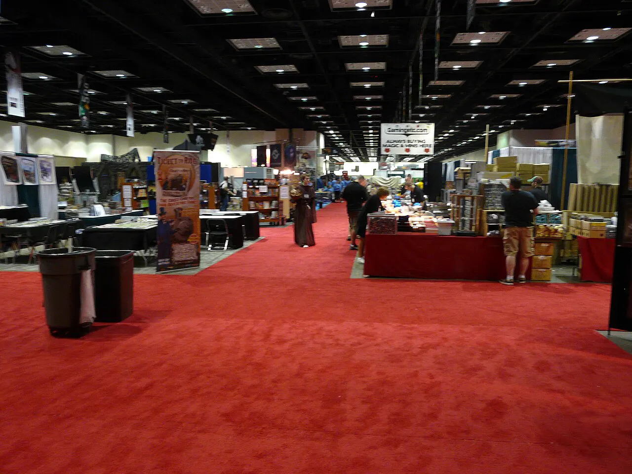 A wide red-carpeted aisle running down the middle of the Gen Con exhibit hall between draped vendor booths, under a black-painted ceiling grid hung with stage lights, with only a handful of people standing at booths in the distance and two black trash bins in the near foreground.
