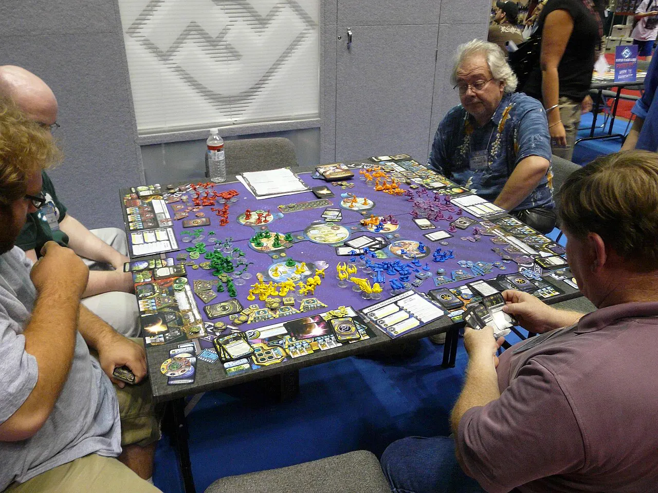 Four players seated around a convention table at Gen Con playing a large hex-map board game covered in red, green, yellow and purple plastic figures, with card trays and player boards ringing the edge of the board and one player holding a fan of cards.