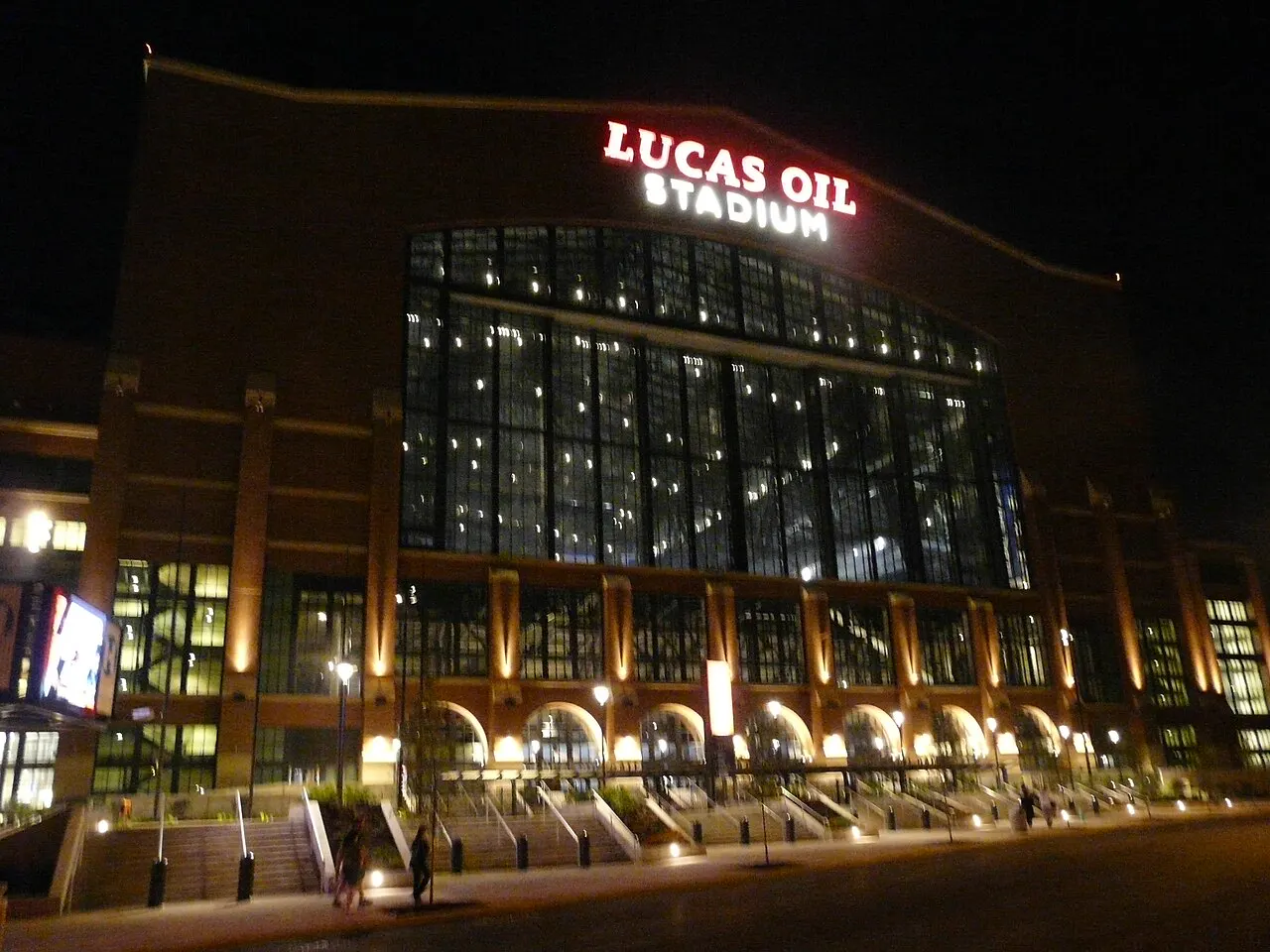 The tall glass west facade of Lucas Oil Stadium in downtown Indianapolis lit from within at night, with the red LUCAS OIL STADIUM sign glowing above the window wall and the empty plaza steps and streetlights below.