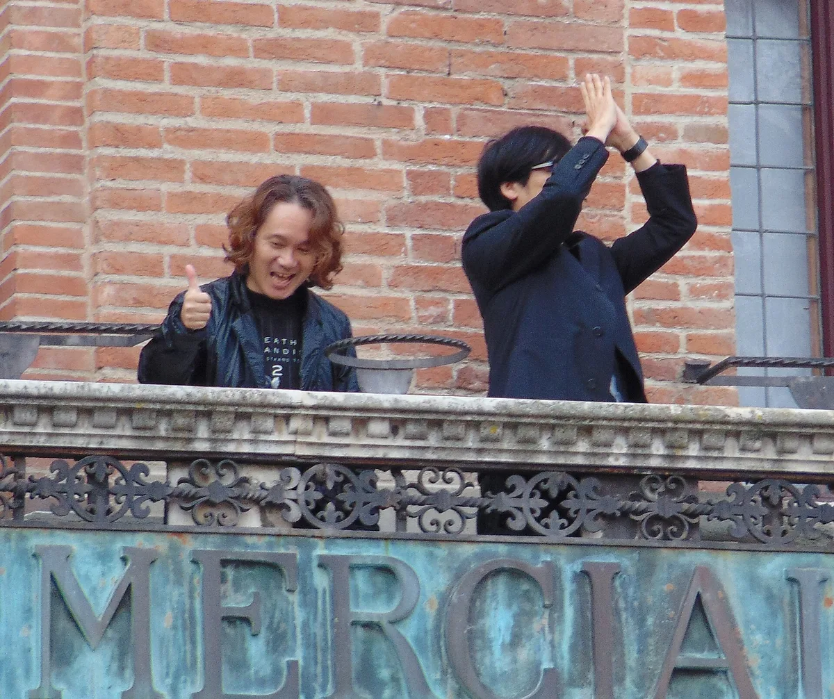 Yoji Shinkawa, with shoulder-length wavy hair and a T-shirt with Death Stranding 2 lettering, grins and gives a thumbs-up beside Hideo Kojima, who raises both hands above his head, on a stone balcony with an ornate iron railing in front of a red brick wall during Lucca Comics & Games 2025. Large weathered green-bronze letters run along the front of the balcony.