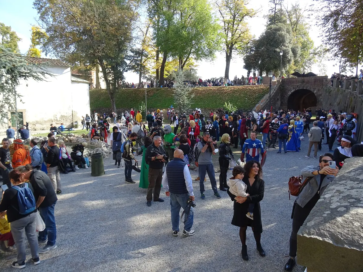 A sunny gravel square at the foot of Lucca's tree-lined city walls during Lucca Comics & Games 2021, packed with cosplayers and visitors: a Captain America cosplayer with his shield stands near the centre, a woman holds a toddler, a man takes a selfie, photographers aim cameras, a small rock-edged pond sits on the left beside a white building, and more people line the top of the grassy rampart near a stone archway on the right.