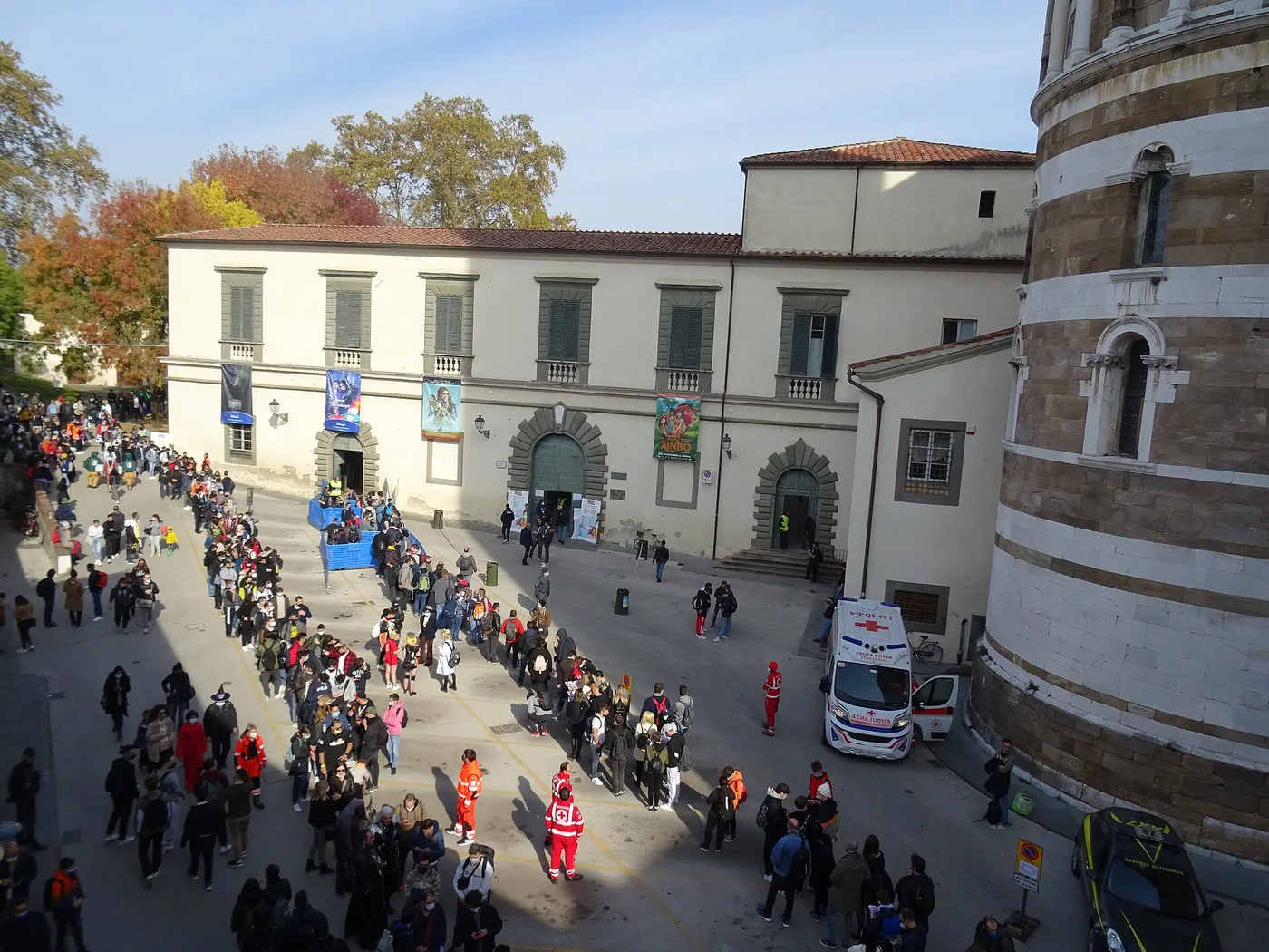 A long queue of visitors, many with backpacks, snakes across a sunny paved square towards the pale facade of Lucca's Real Collegio, hung with film banners, during Lucca Comics & Games 2021. Blue crowd barriers stand near the doorway, Red Cross volunteers in red and orange uniforms stand in the foreground, and an ambulance is parked beside a striped stone building on the right.