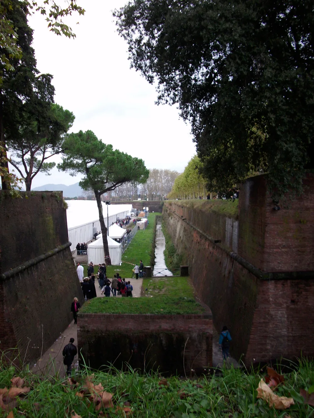 A long white exhibition tent stretches along the grassy moat below Lucca's brick city walls during Lucca Comics & Games 2008, photographed from the ramparts, with smaller white gazebos, crowd barriers and visitors on the path beside it and hills in the distance.