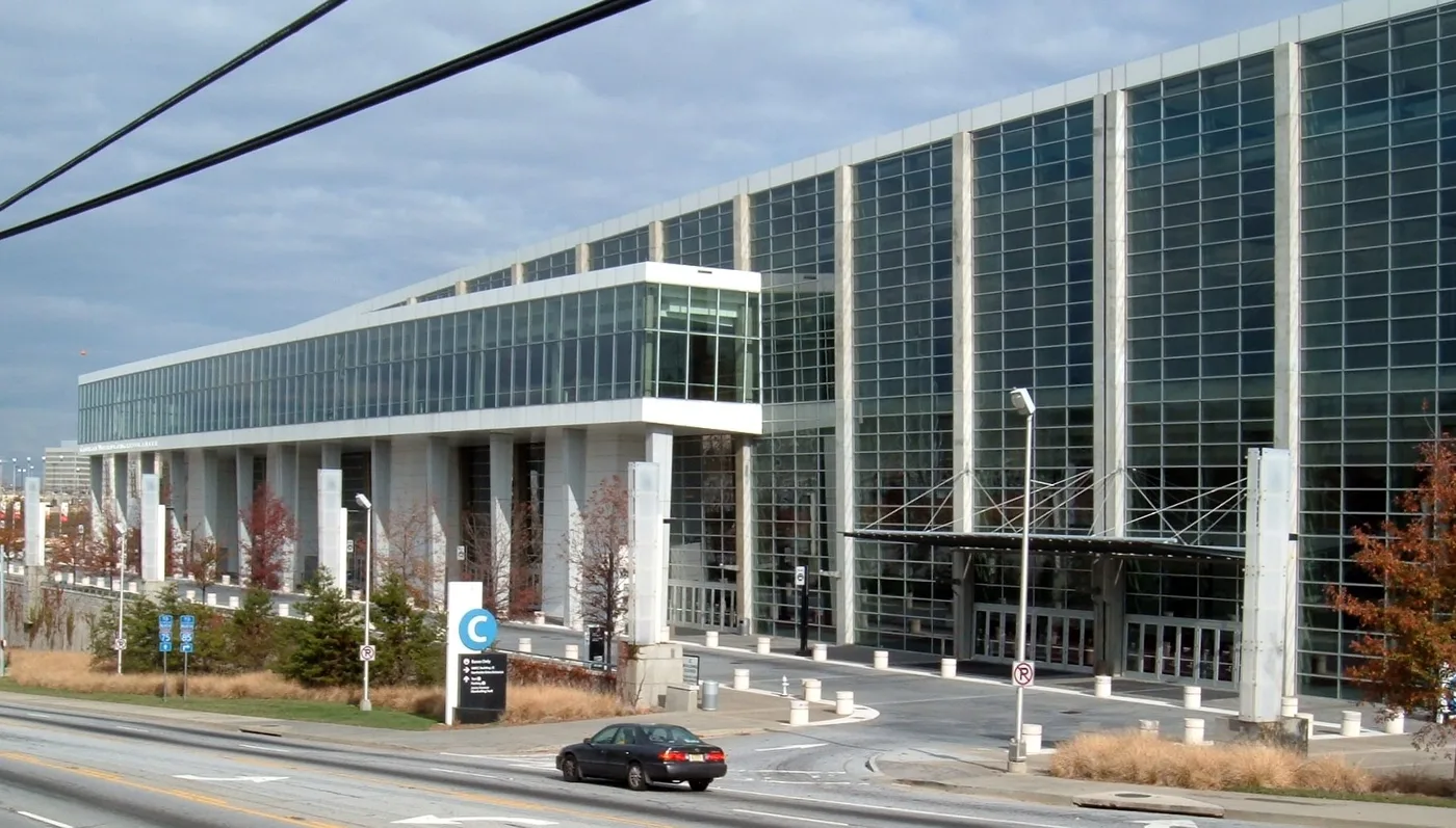 The Georgia World Congress Center seen from across Northside Avenue, a long white-columned building with a two-storey glass curtain wall, a covered entrance canopy and a large blue letter C wayfinding sign at the kerb.