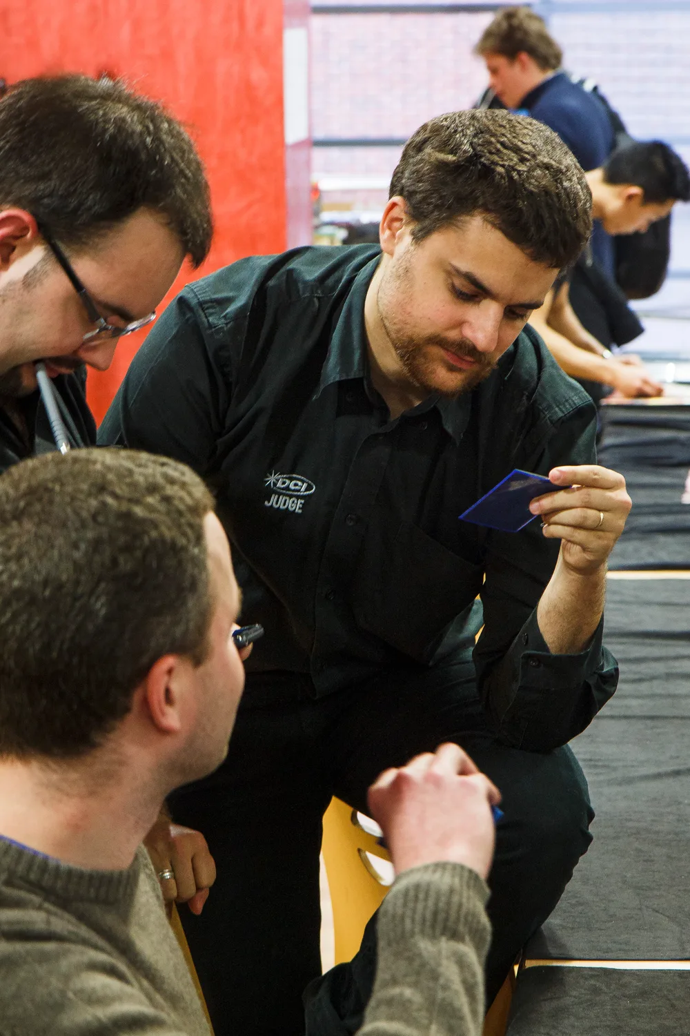 A DCI-shirted Magic judge crouching beside a tournament table, holding a card up and reading it closely while two seated players in the foreground watch and wait for the ruling.