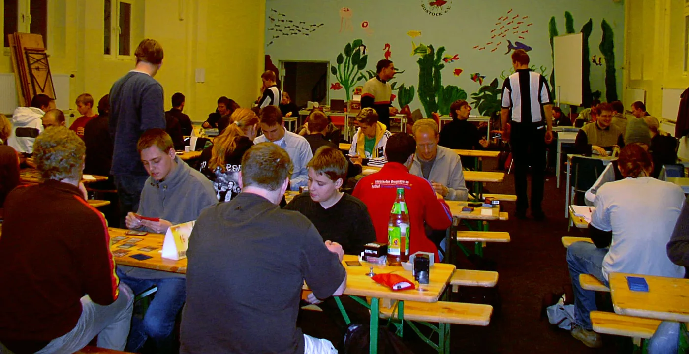 A hall of long wooden tables packed with Magic players seated opposite each other mid-game, cards and dice spread out, with a standing judge in a striped shirt watching over the room.