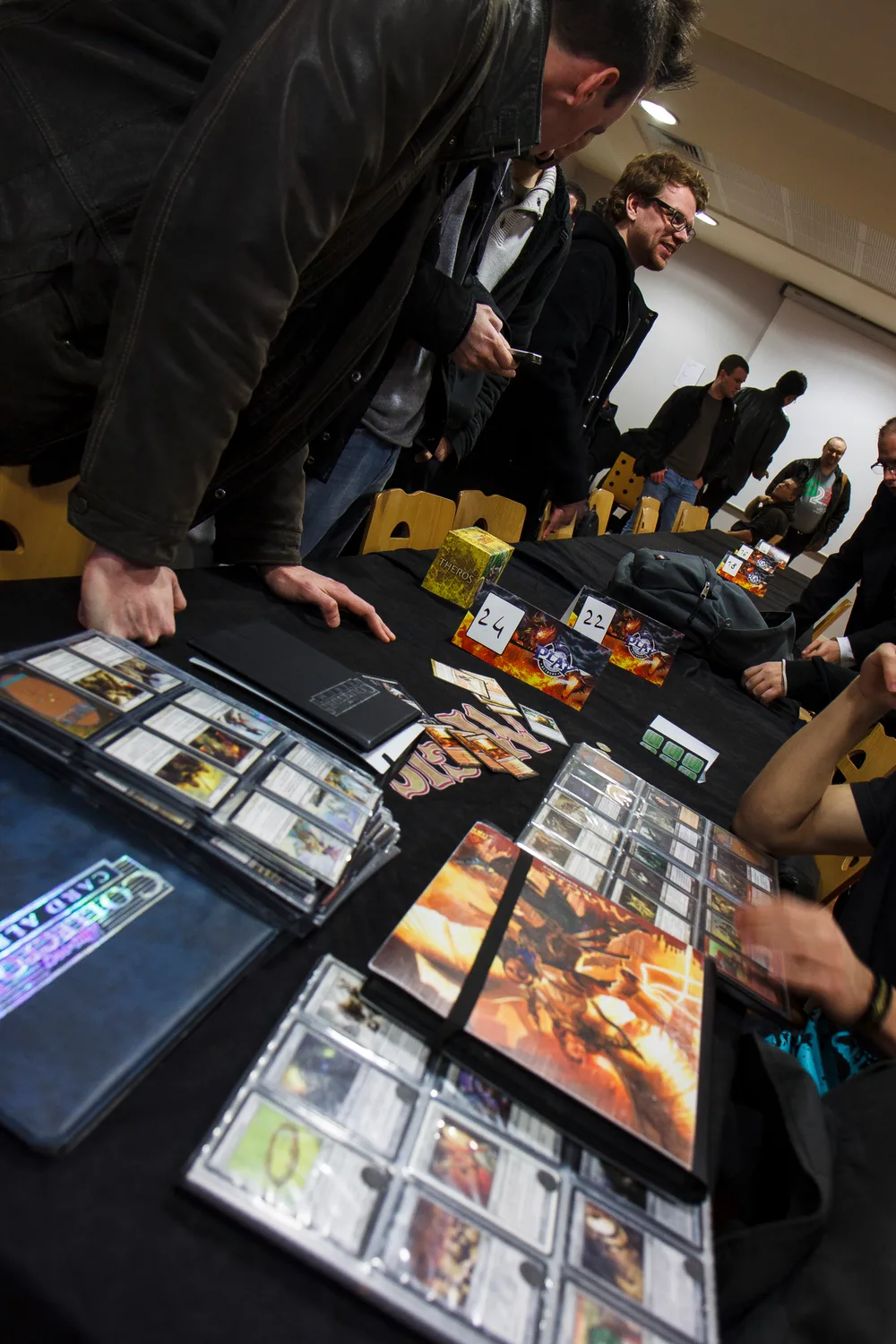 Magic players leaning over a convention table covered in open card binders, loose cards, a boxed Theros set and numbered table placards, trading and browsing during a tournament.