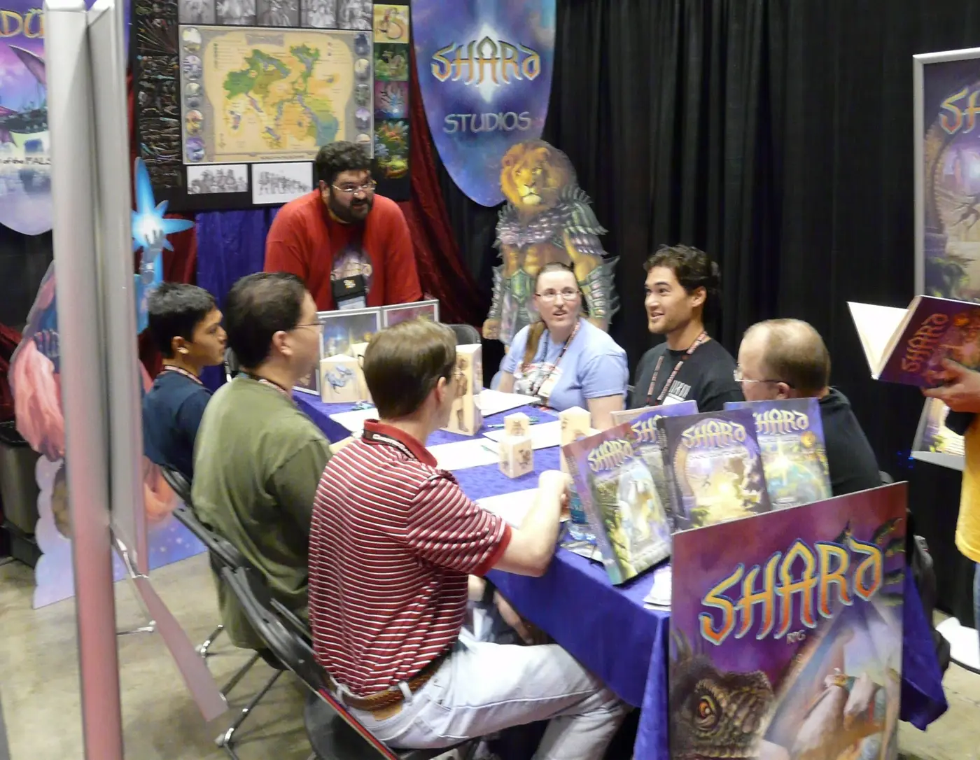 A publisher booth at Gen Con Indy: a bearded staffer in a red shirt stands behind a purple-draped table explaining a game to five seated attendees wearing convention lanyards, with boxed Shara RPG product propped upright along the table edge and a Shara Studios banner hanging above the booth.