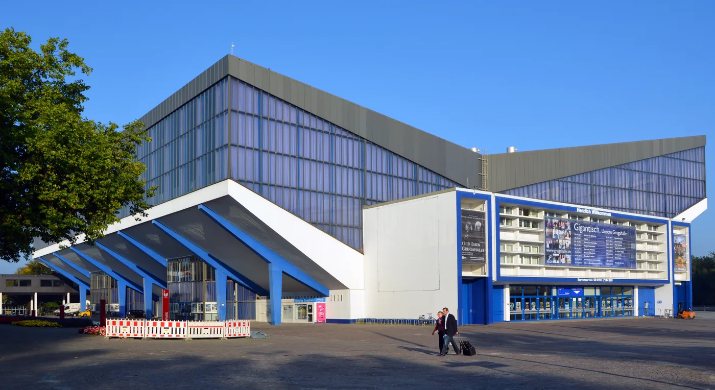 The Grugahalle in Essen on a clear day: a long hall with a steeply sloping blue glass facade and a jutting white-and-blue cantilevered entrance canopy, a large advertising banner on the right-hand wing and two people wheeling a suitcase across the paved forecourt.