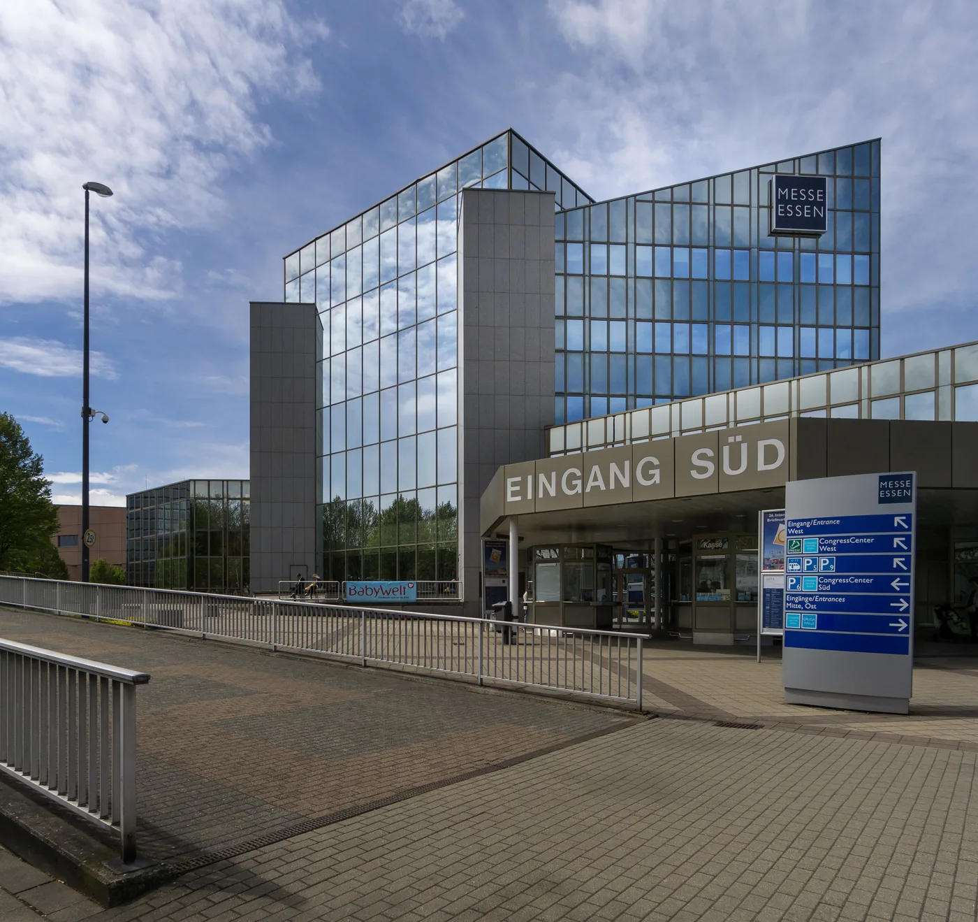 The Entrance South building at Messe Essen: a tall glass-and-steel tower with a MESSE ESSEN sign, a low canopy lettered EINGANG SÜD over the doors, and a blue wayfinding sign in the foreground pointing to the West and East entrances and the Congress Center.