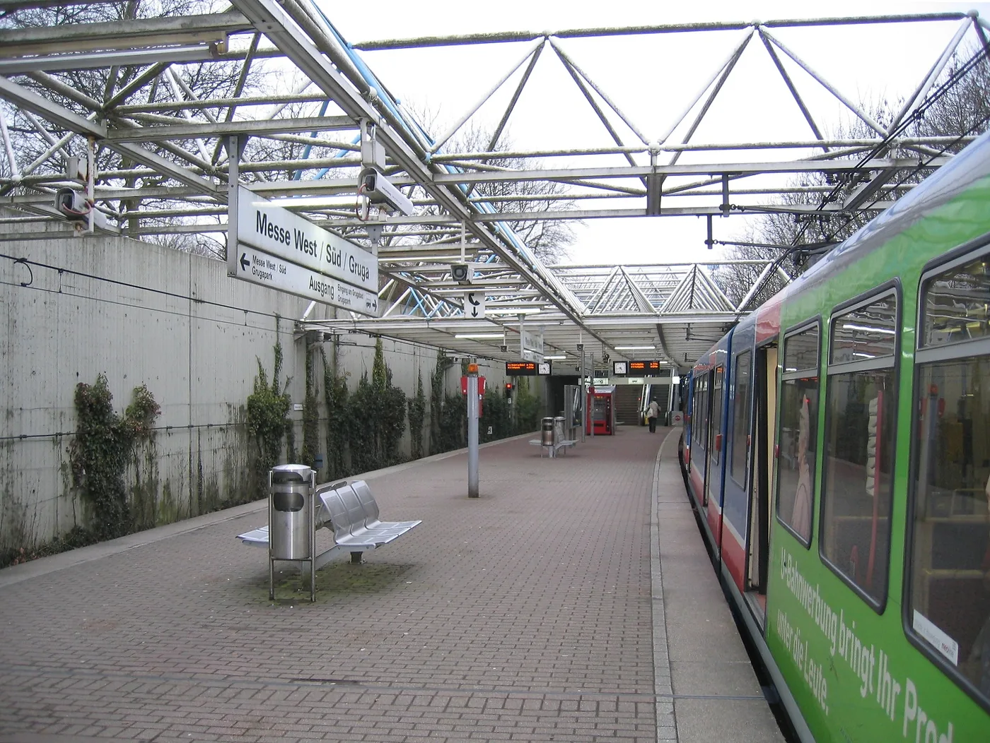 The Messe West/Süd/Gruga U-Bahn platform in Essen: a long brick-paved platform under a white steel truss canopy, an overhead sign reading Messe West / Süd / Gruga, benches and a bin in the foreground, and a green-liveried tram waiting on the right.
