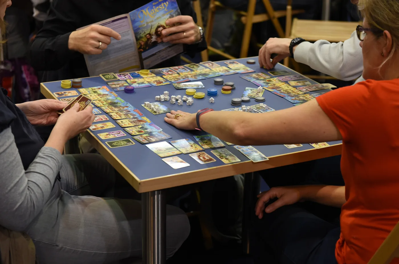 Four players around a small table at SPIEL 2017 in Essen, one holding the Majesty rulebook open while the others play building cards, coloured chips and small white pieces on a blue play mat.