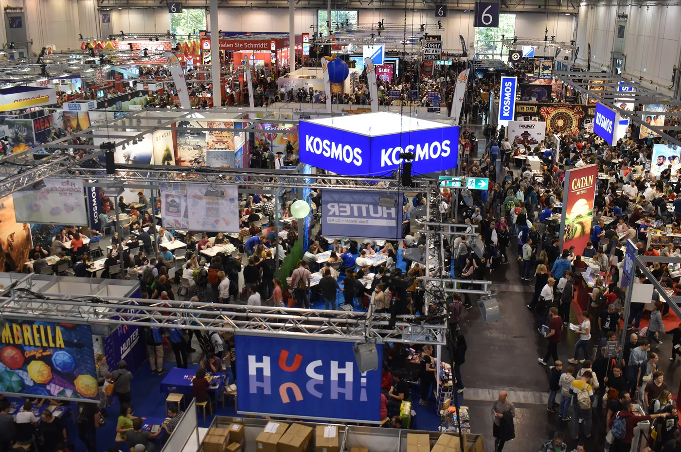 Hall 6 at SPIEL Essen 2024 seen from above: crowds walk the aisles and sit at demo tables between publisher booths, with a large blue KOSMOS banner cube in the centre, HUCH and HUTTER signs below it, a CATAN banner on the right and a hall marker reading 6 hanging from the roof.