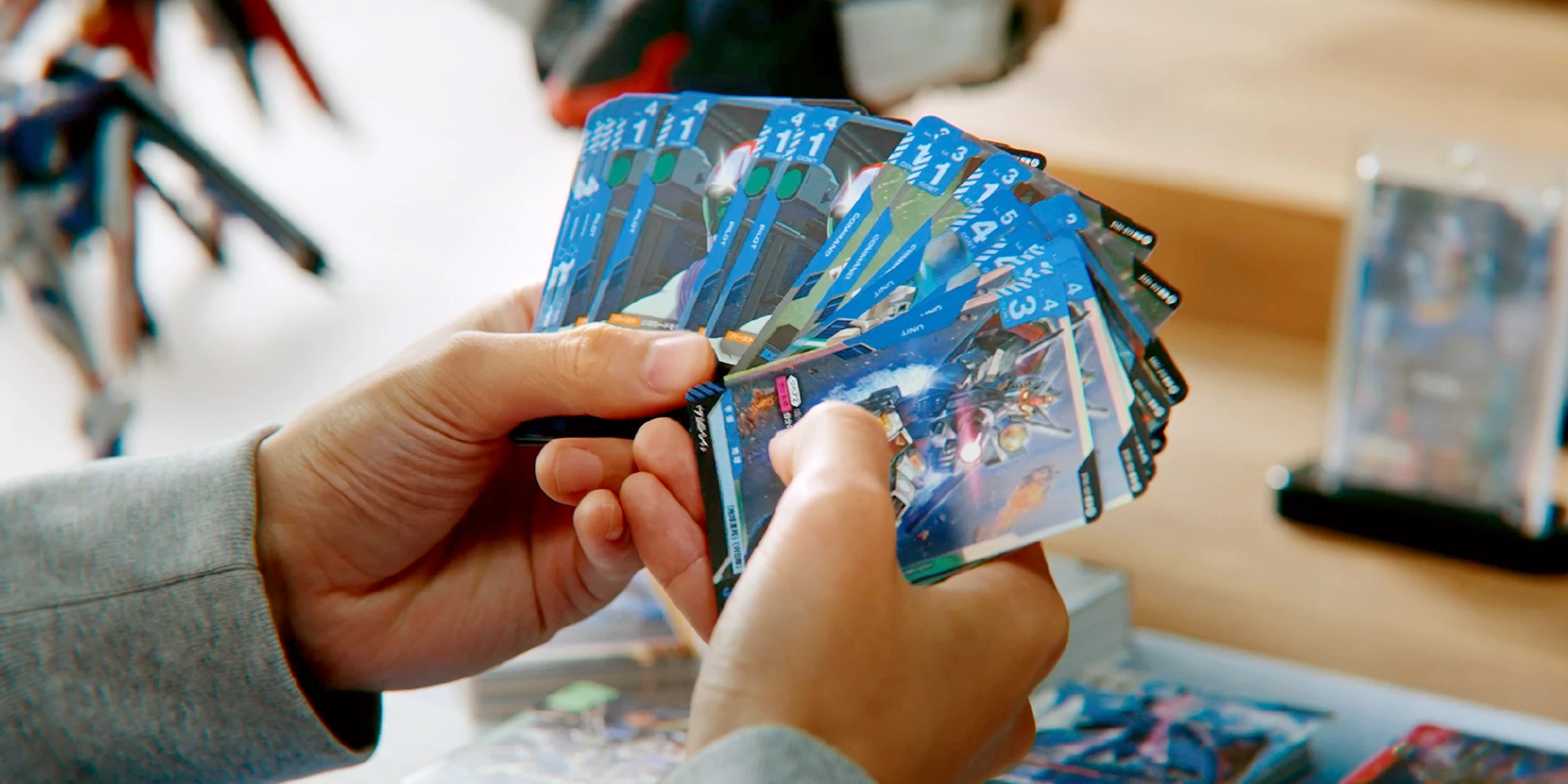 A player holding a fanned-out handful of blue Gundam Card Game cards over a wooden table, with more cards and a mobile suit model kit in the background.