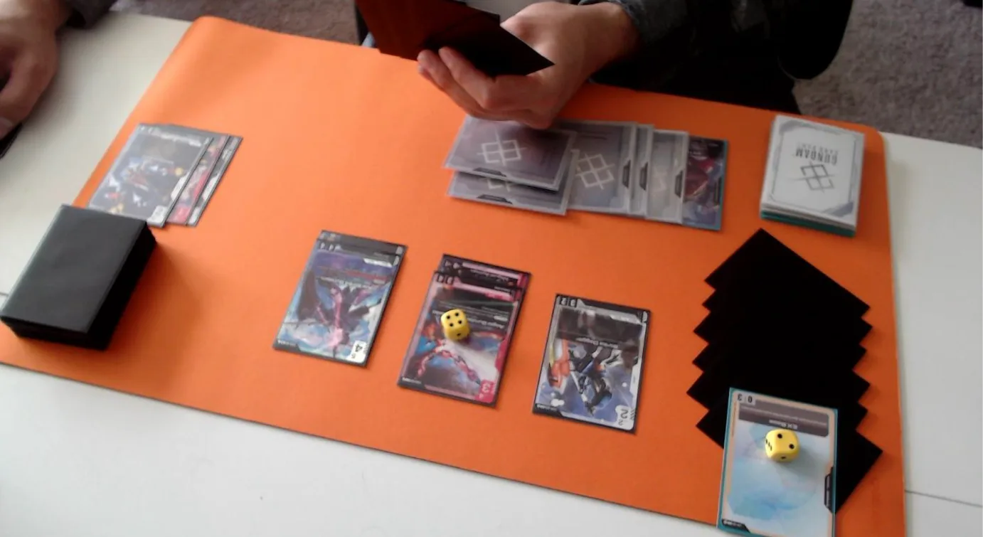 A photo from Bandai's Tournament Rules Manual showing a correct webcam setup on an orange playmat: a hand of black-sleeved cards held up with its backs to the camera, a black-sleeved deck and a row of face-up cards on the left, three Units in the centre with a yellow die on the middle one, a row of clear-sleeved cards and a stack with the Gundam Card Game card back along the top, black-sleeved Shields fanned out on the right so each one can be counted, and an EX Base with a yellow die on it at the front of the shield area.
