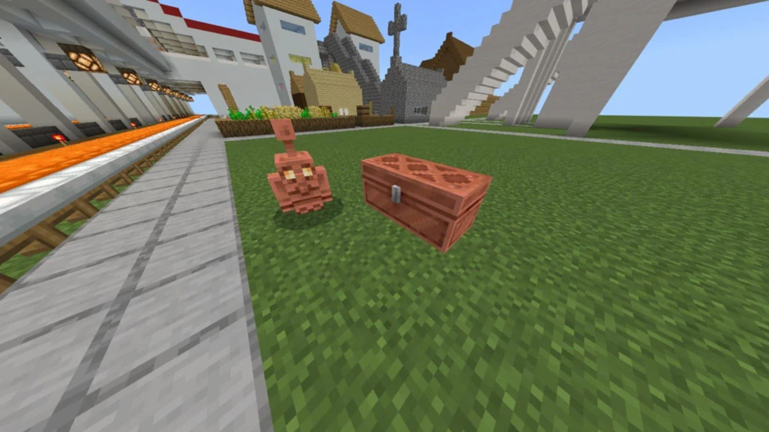 A copper golem standing next to a copper chest on a grass field, with a redstone build and village houses behind it.