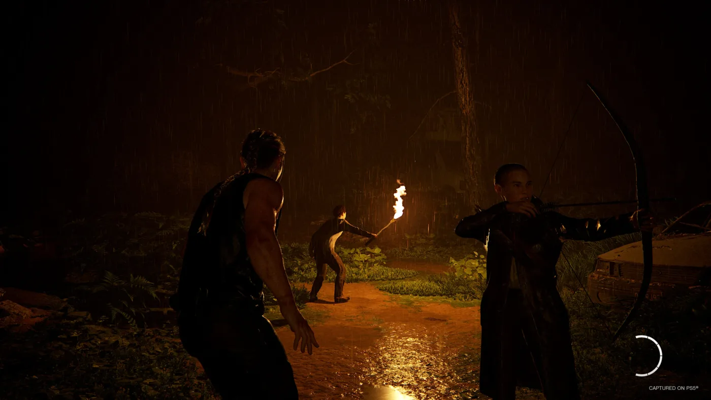 Abby, in a sleeveless top with her braid down her back, squares up beside Lev as he draws a bow on a rain-soaked forest road at night in The Last of Us Part II Remastered, while a figure holding a lit torch stands on the path ahead of them.
