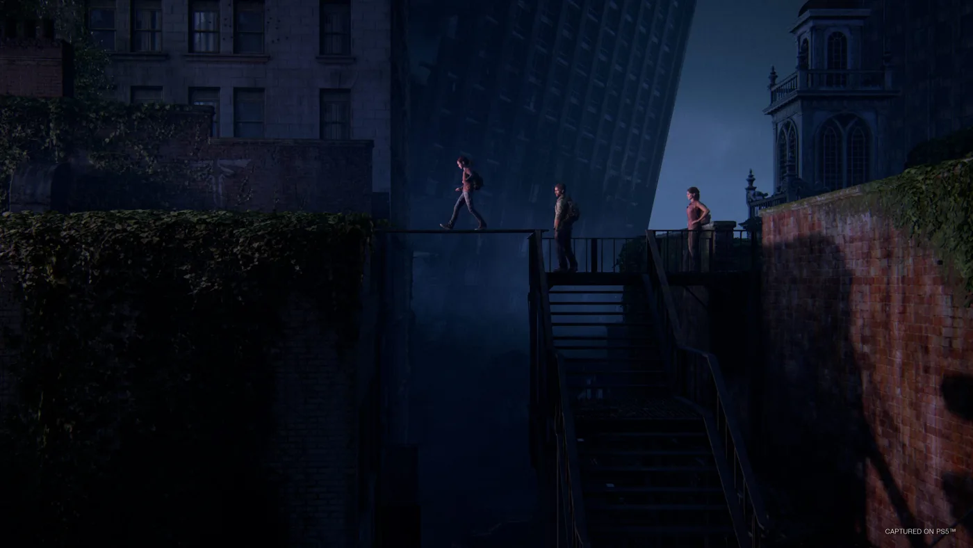 Ellie, Joel and Tess crossing a narrow metal walkway between ivy-covered buildings in the Boston quarantine zone in The Last of Us Part I.